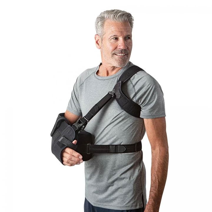 Man wearing a black arm sling and waist support belt over a gray T-shirt, standing against a white background