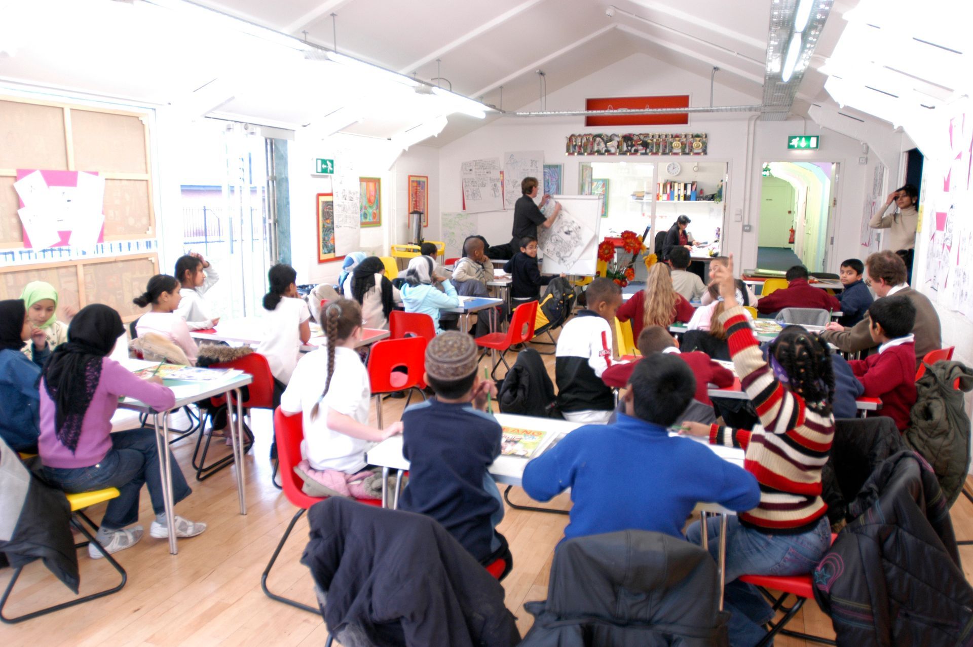 Children in a brightly lit art class. Students sit at tables, some raise hands. A teacher stands at a whiteboard.