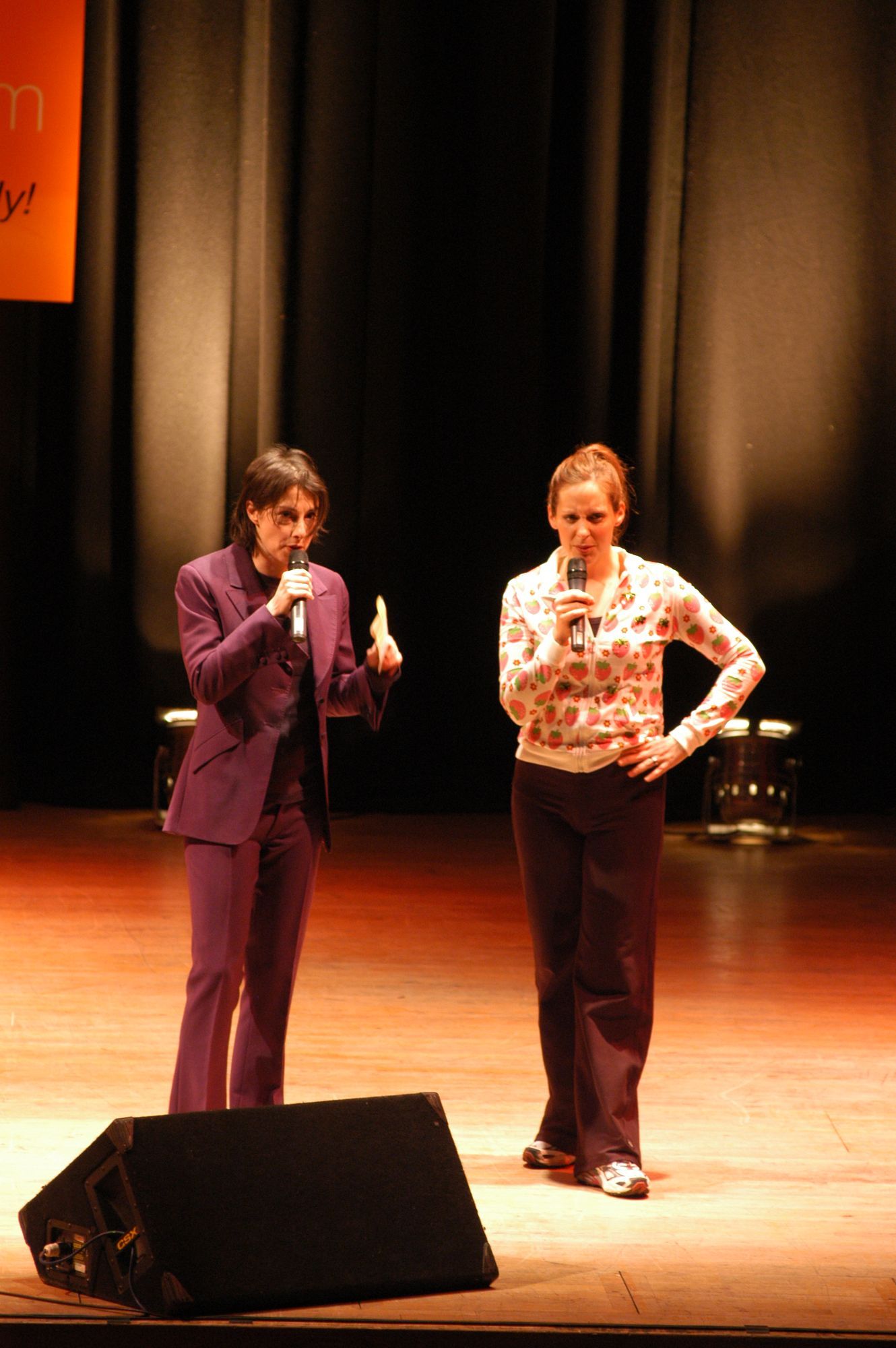 Two people on stage with microphones. One in purple suit gestures, the other in print shirt with hand on hip.