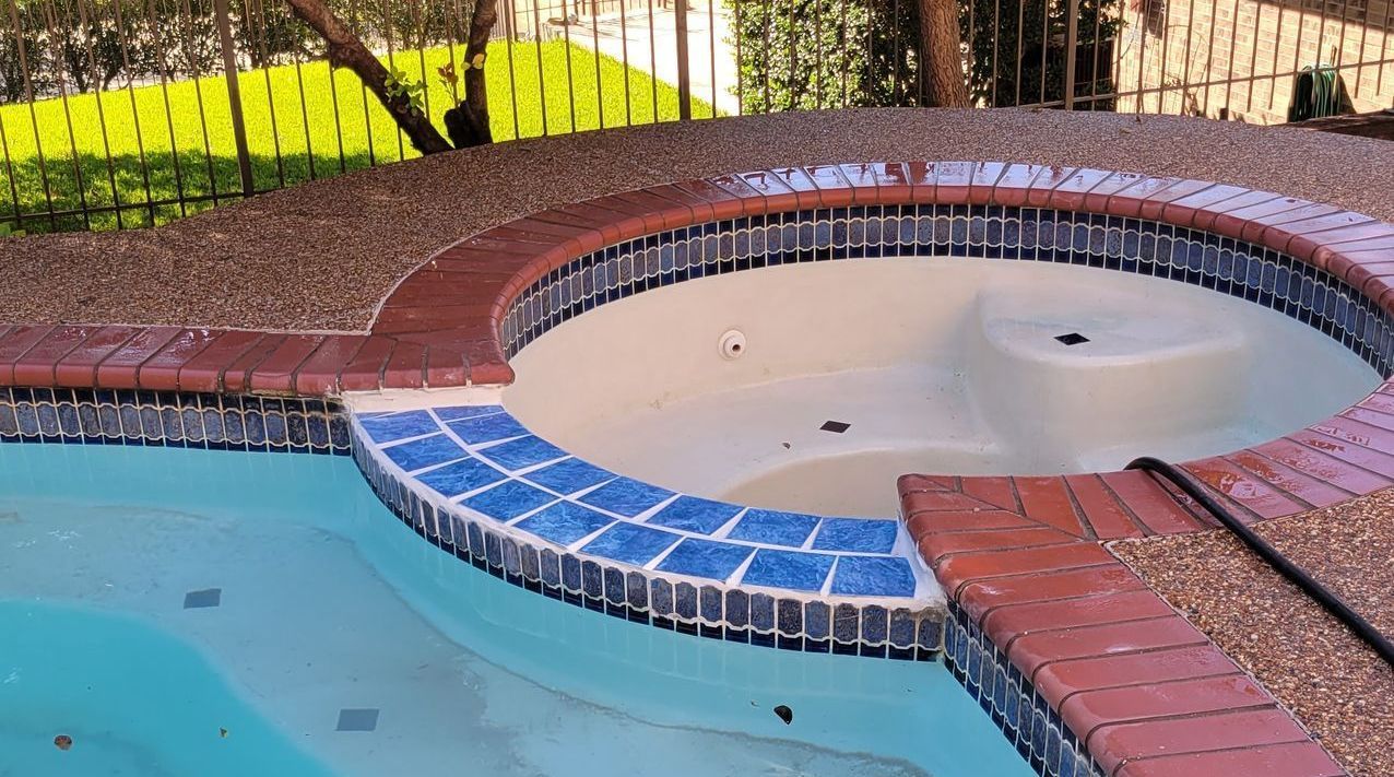 A round hot tub with a light interior and blue tile trim sits next to a pool with light blue water and a brick border.