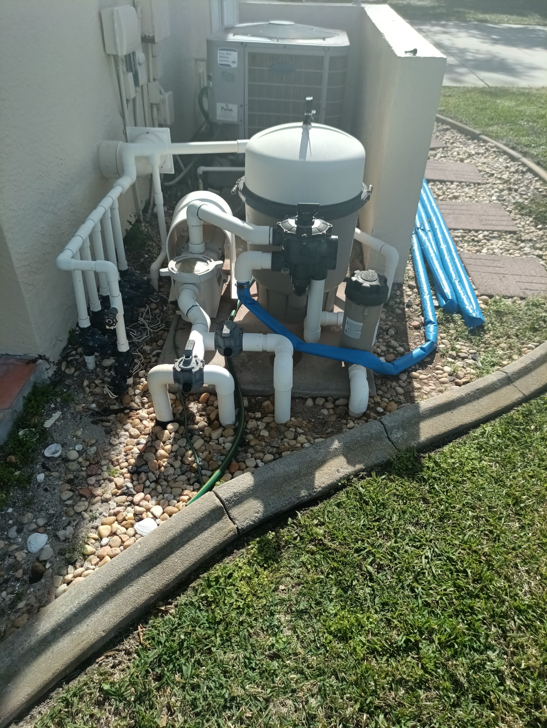 Old Pool filter and pump equipment on a gravel base next to a building and lawn.