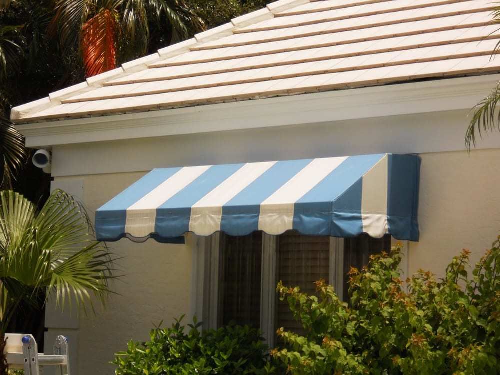 A blue and white striped window awning attached to the exterior wall of a white house surrounded by green foliage.
