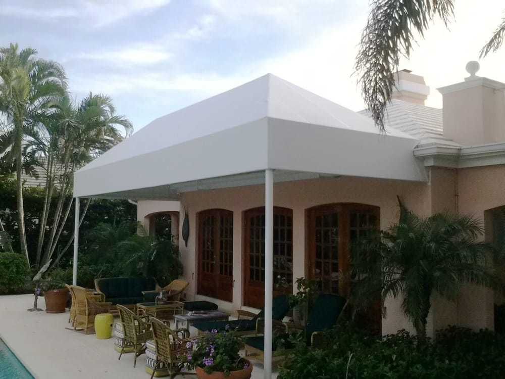 A white fabric awning covers a patio area with outdoor furniture next to a light-colored house and a swimming pool.