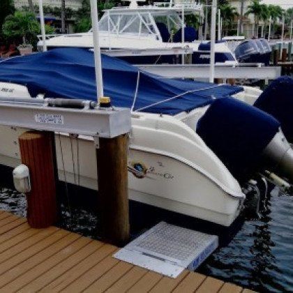 A white boat with a blue cover is docked at a wooden pier, with another boat visible in the background.