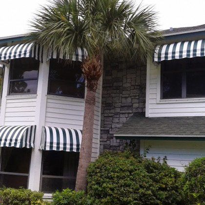 A two-story house with white horizontal siding, stone accents, and green and white striped awnings over the windows.