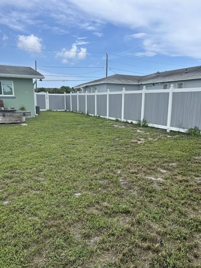 A backyard with patchy green grass, a green house on the left, and a white privacy fence spanning the back.