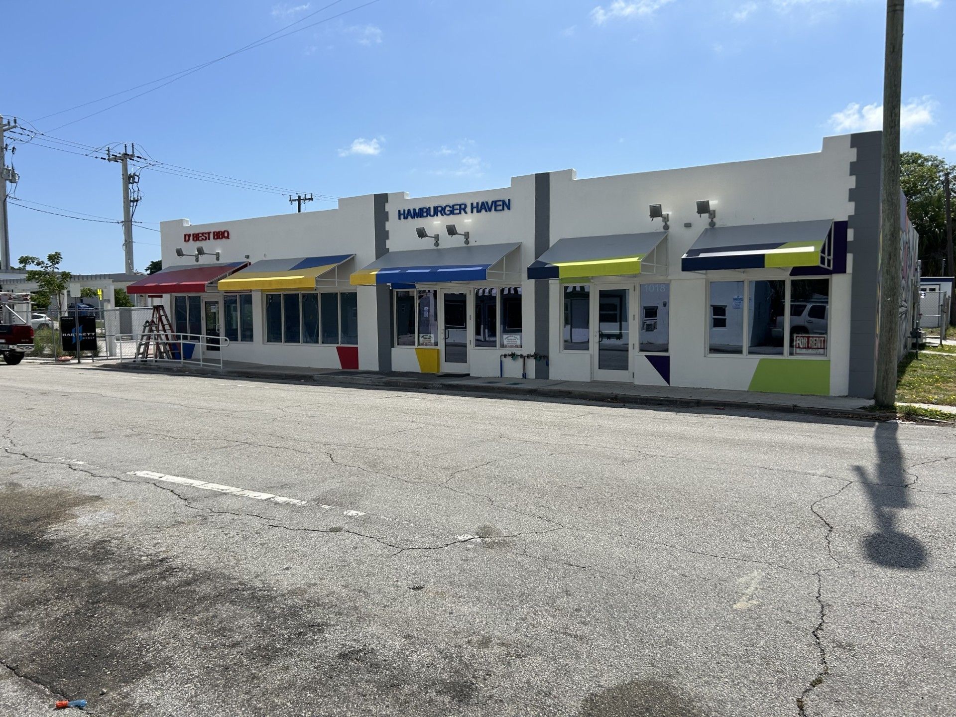 A one-story commercial strip mall building with colorful awnings along the front, situated beside a paved road.