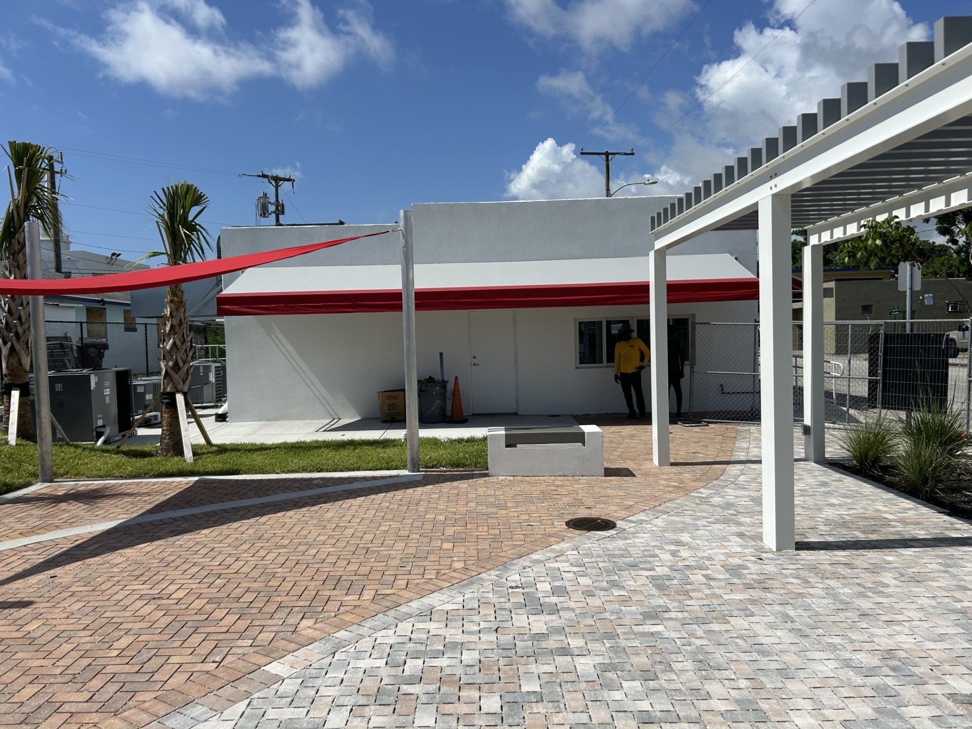 A white building under a red awning, featuring a red shade sail and a white pergola over a brick-paved patio area.