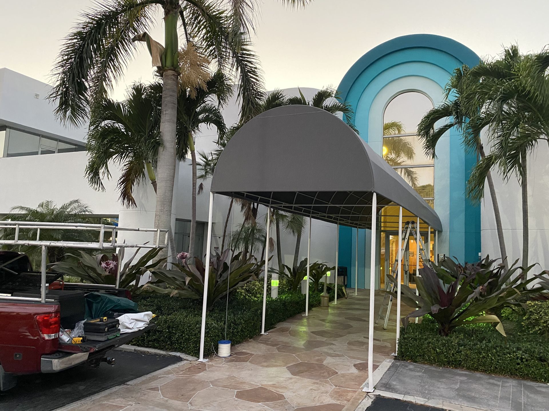 A covered walkway leads to a building entrance with a blue arched frame, surrounded by palm trees and a parked truck.
