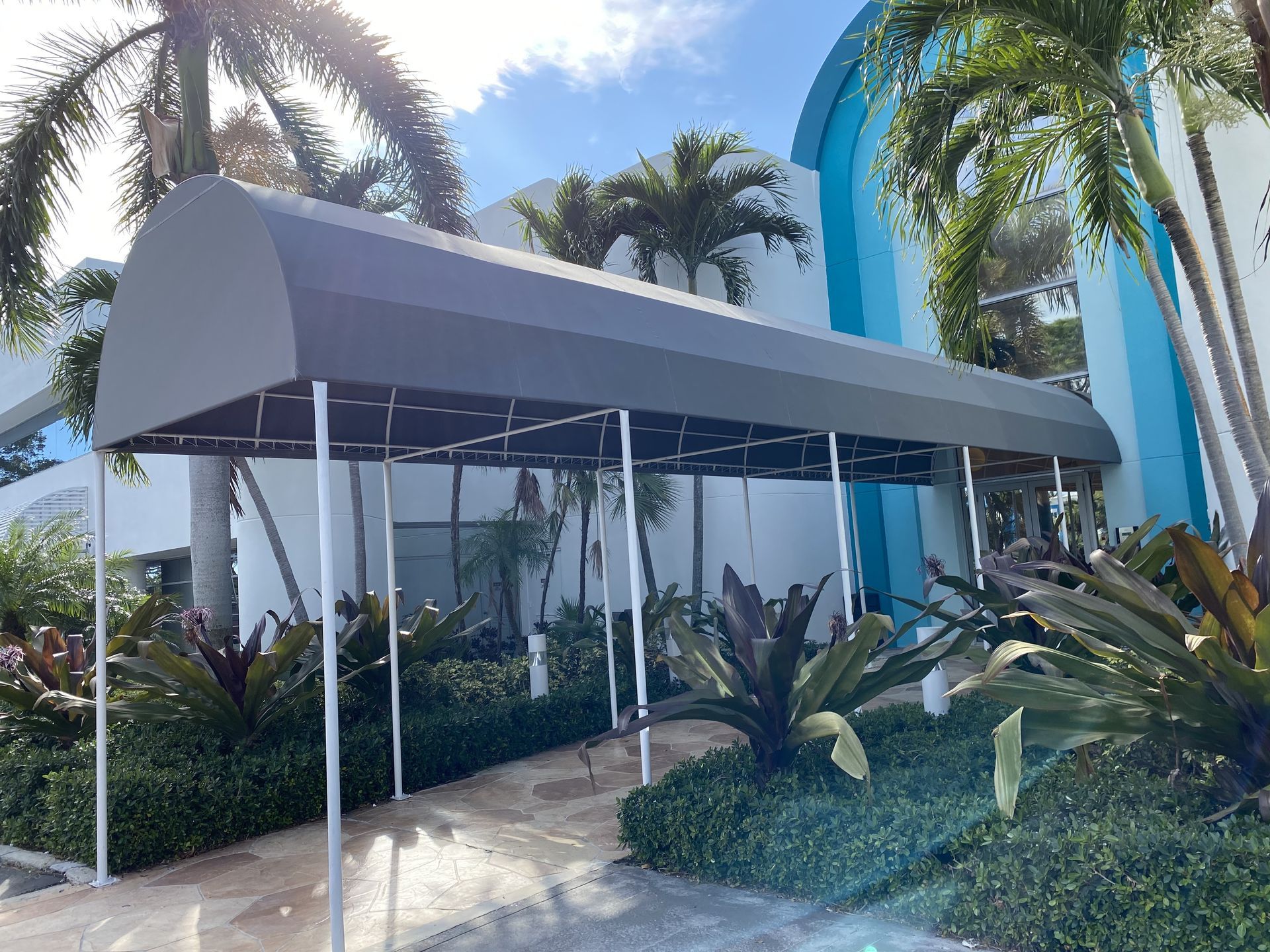 A long gray arched canopy walkway leads to the blue-trimmed glass entrance of a building, surrounded by palm trees.