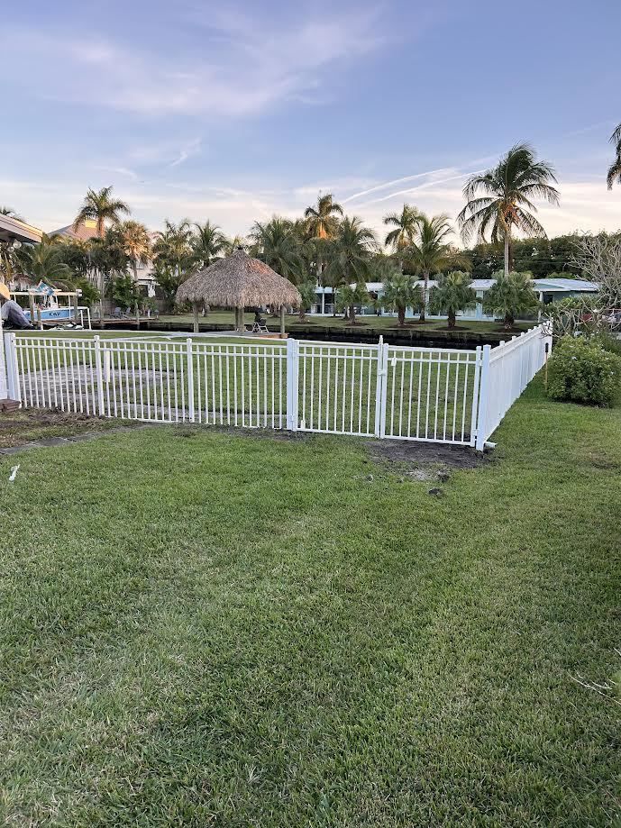 A white picket fence encloses a grassy yard with a thatched-roof tiki hut, trees, and a distant waterway under a blue sky.