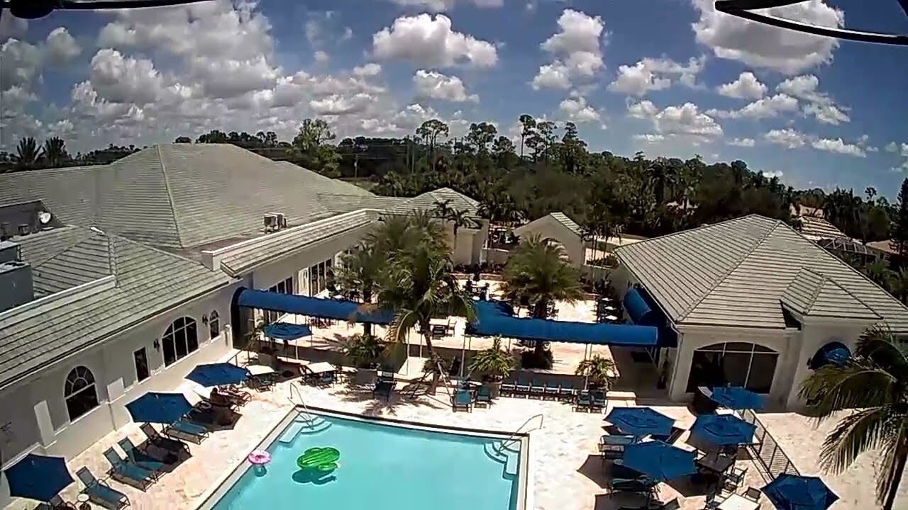 An elevated view of a sunlit resort pool area with beige buildings, blue umbrellas, and a bright green pool float.