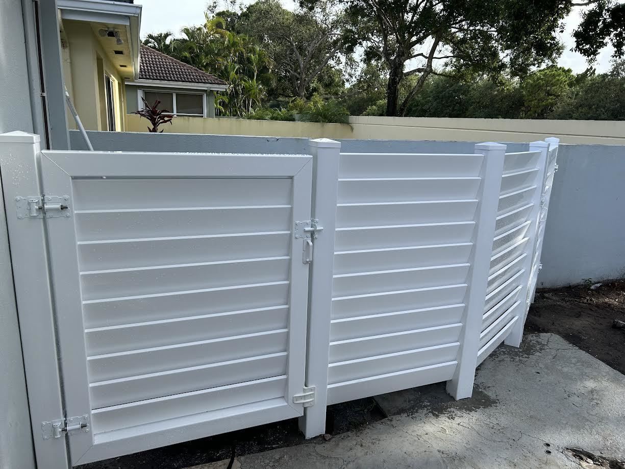 A white, louvered privacy gate and fence section installed on a concrete surface in front of a house.