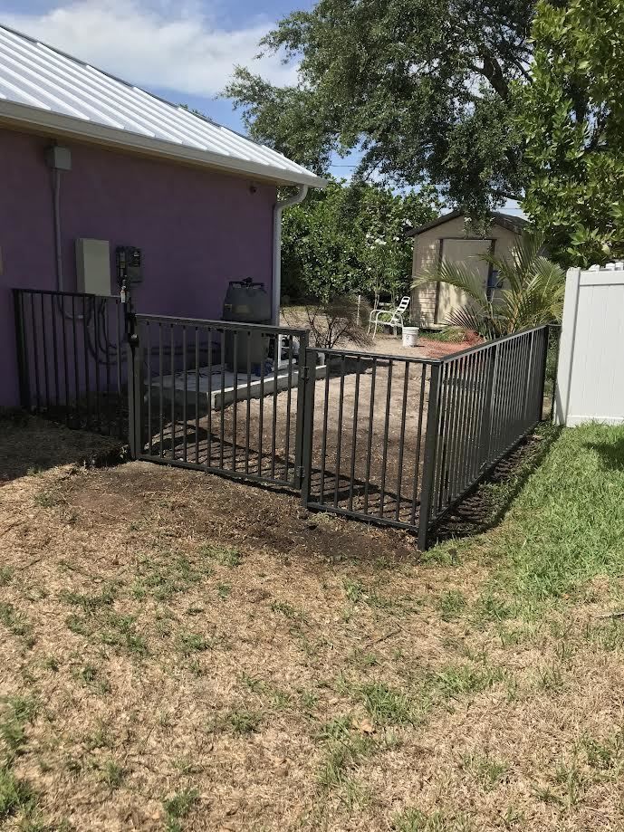 A metal fence encloses a patio area with a grill next to a lavender-colored house in a grassy yard.