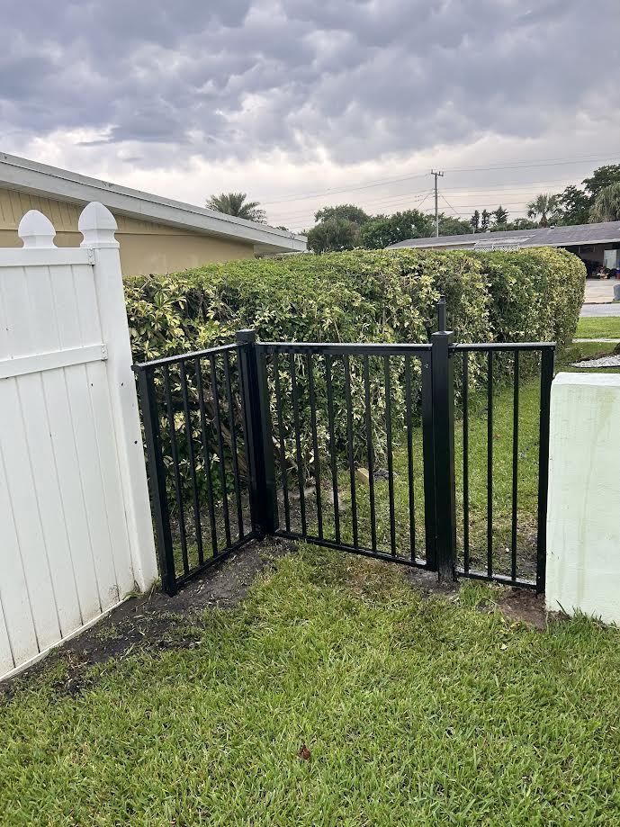 A small black metal gate sits in a grassy yard between a white vinyl fence and a pale green wall, backed by green hedges.