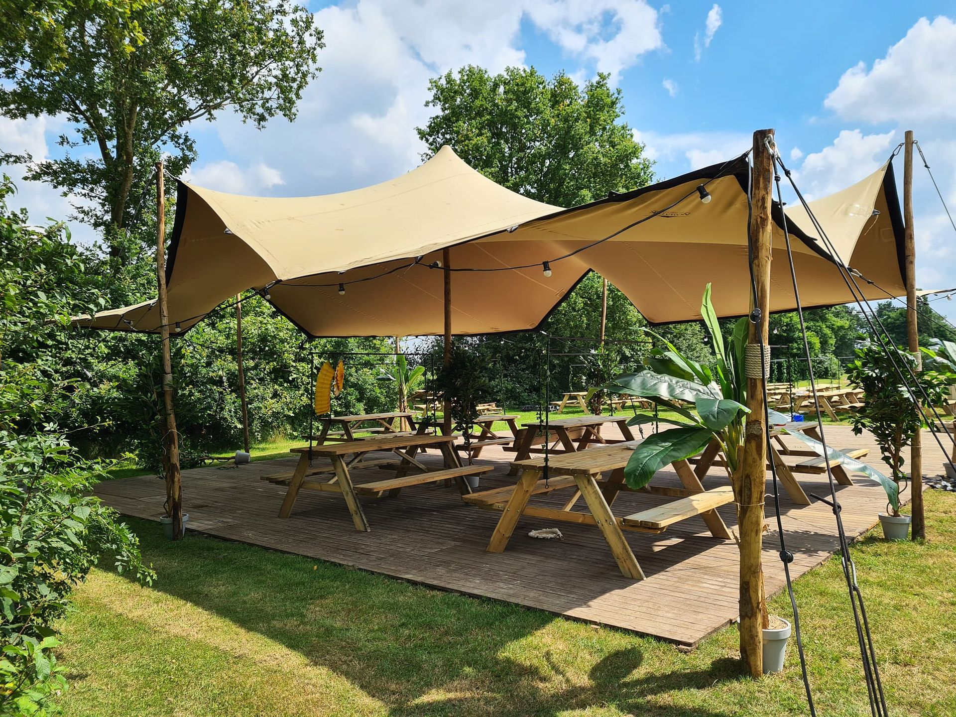Een beige stretchtent biedt schaduw aan picknicktafels op een grindterras; houten palen en lichtslingers.