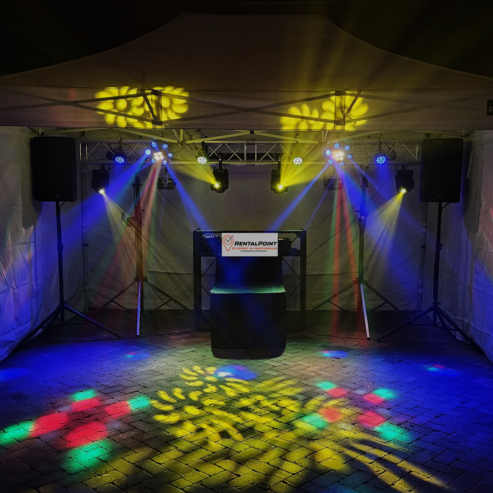 DJ-setup onder een tent met speakers, verlichting en een tafel die verlicht is in geel, blauw, rood en groen.