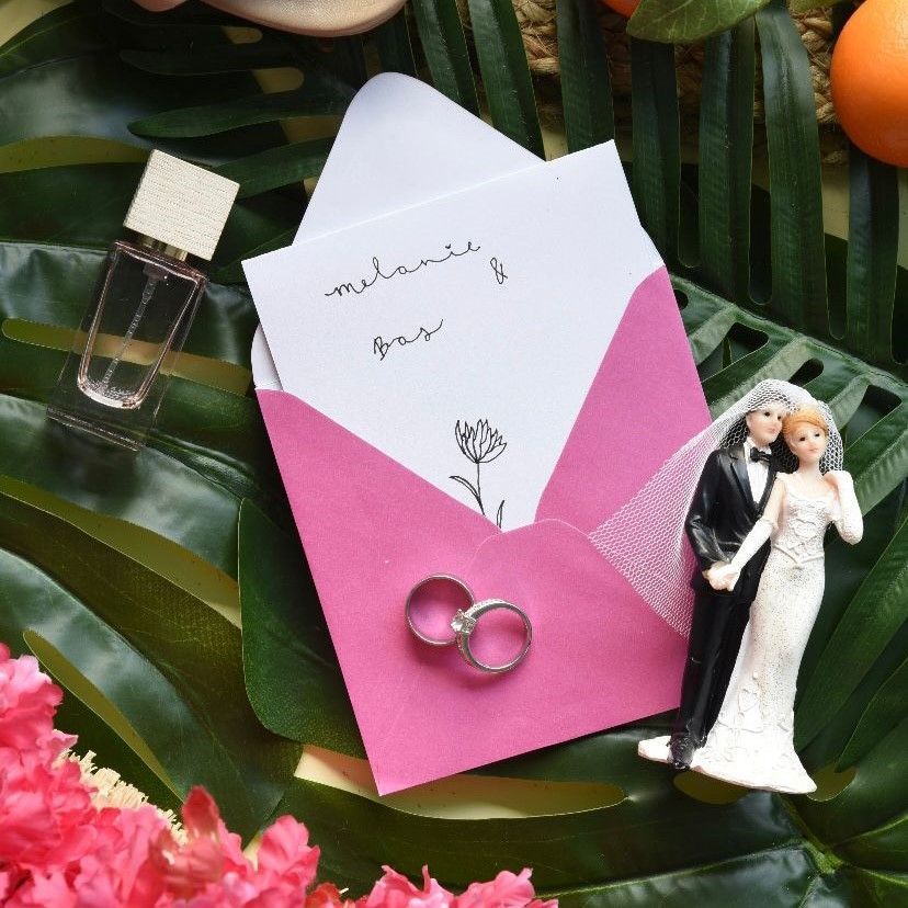 Trouwuitnodiging en ringen op tropisch bladwerk, met parfum en een taarttopper.