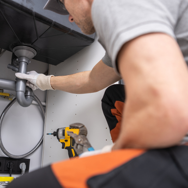 Plumber working under a sink, wearing gloves and orange overalls, using tools.