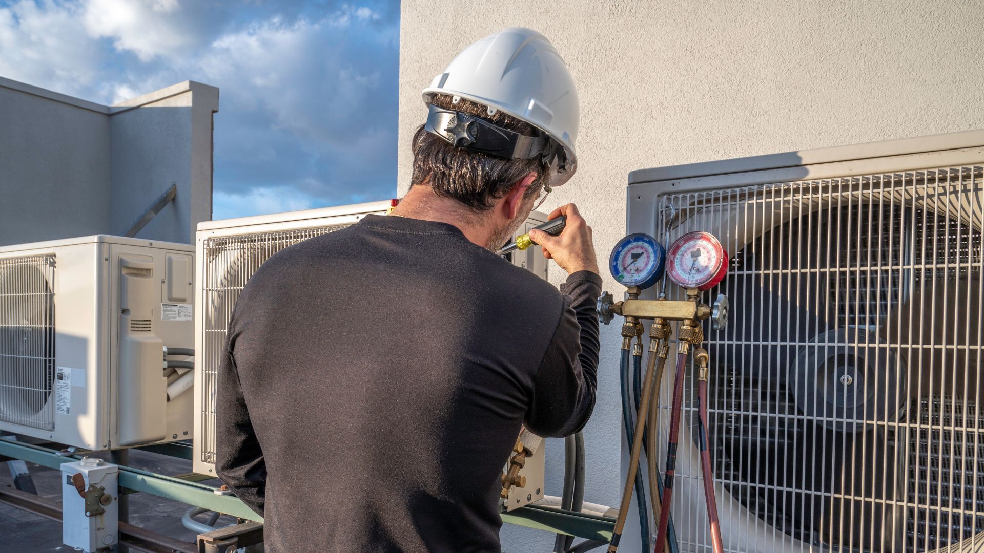 HVAC technician in hard hat, inspecting rooftop air conditioning unit with gauges.