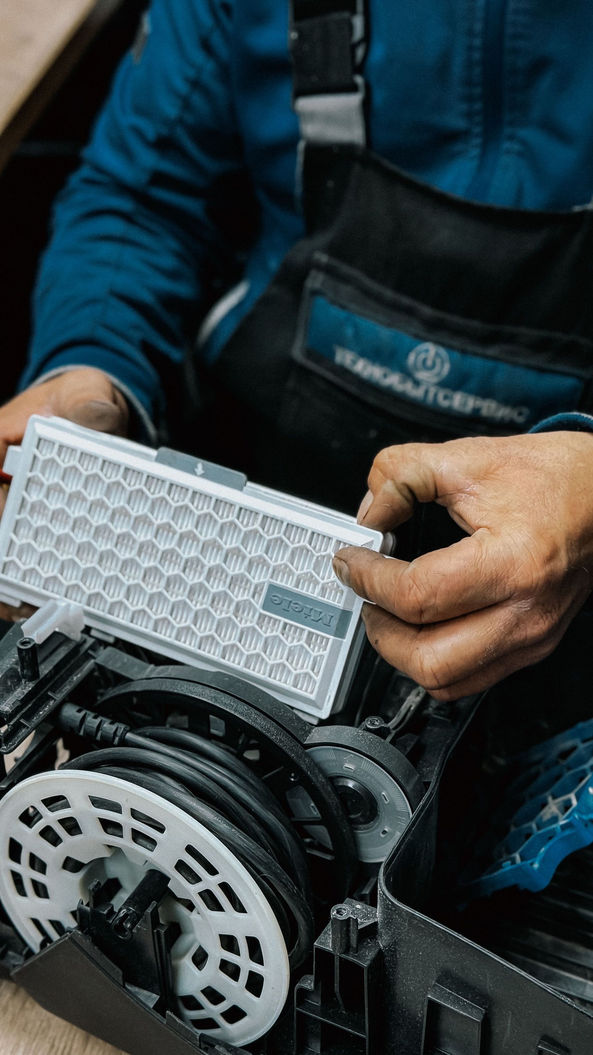 Técnico inspeccionando un filtro de aire de un coche junto al motor, vestido con una chaqueta azul y un delantal negro.