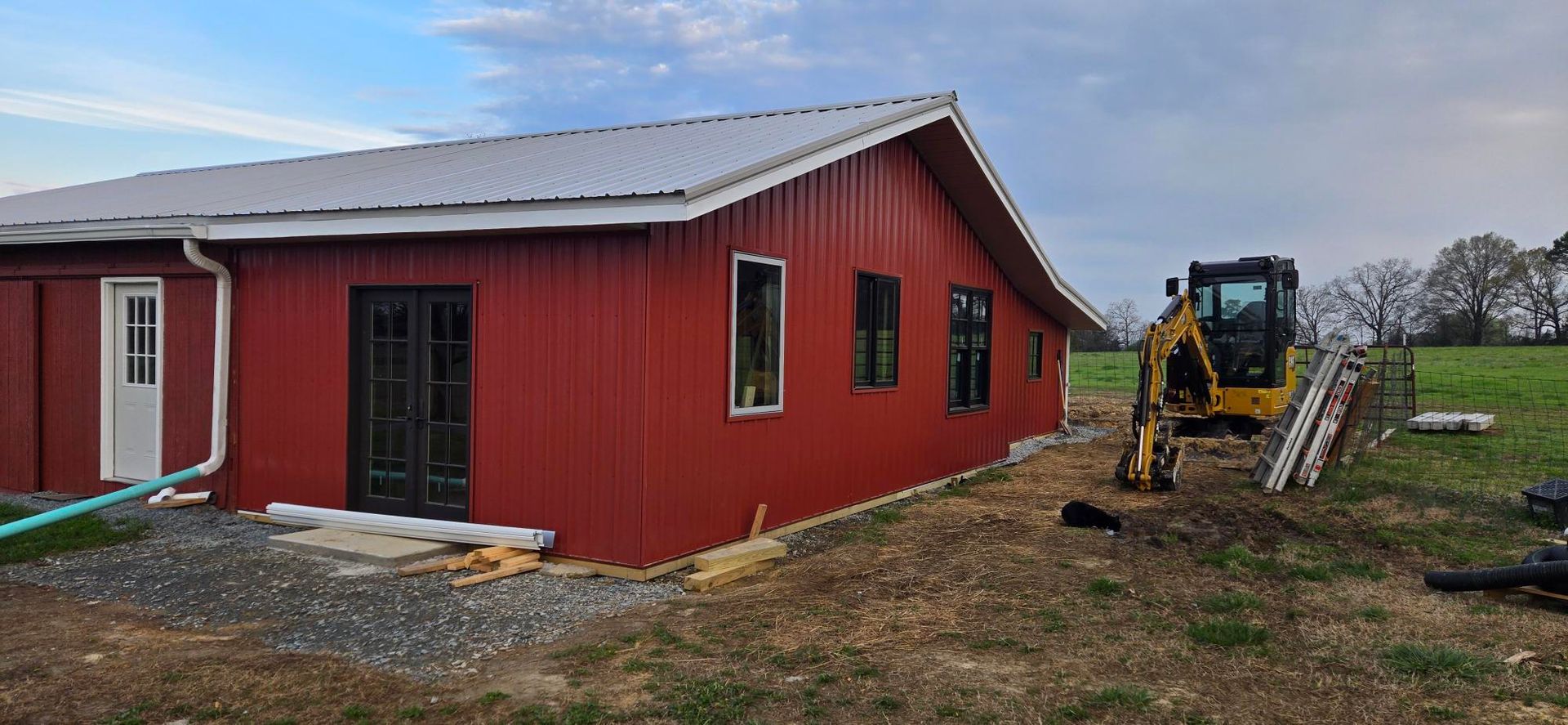 Red building with a metal roof under construction, with a backhoe and ladders on a muddy lot.