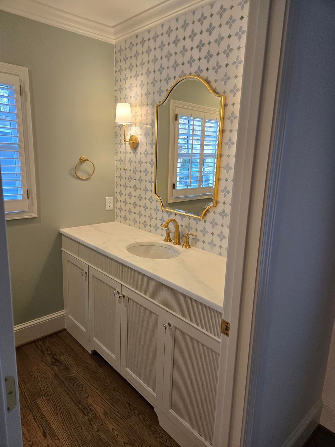White bathroom vanity with gold-framed mirror, sink, wall sconces, and patterned wallpaper next to a window.