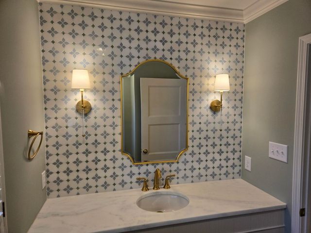 Bathroom vanity with patterned wallpaper, brass mirror, sconces, and a white countertop sink