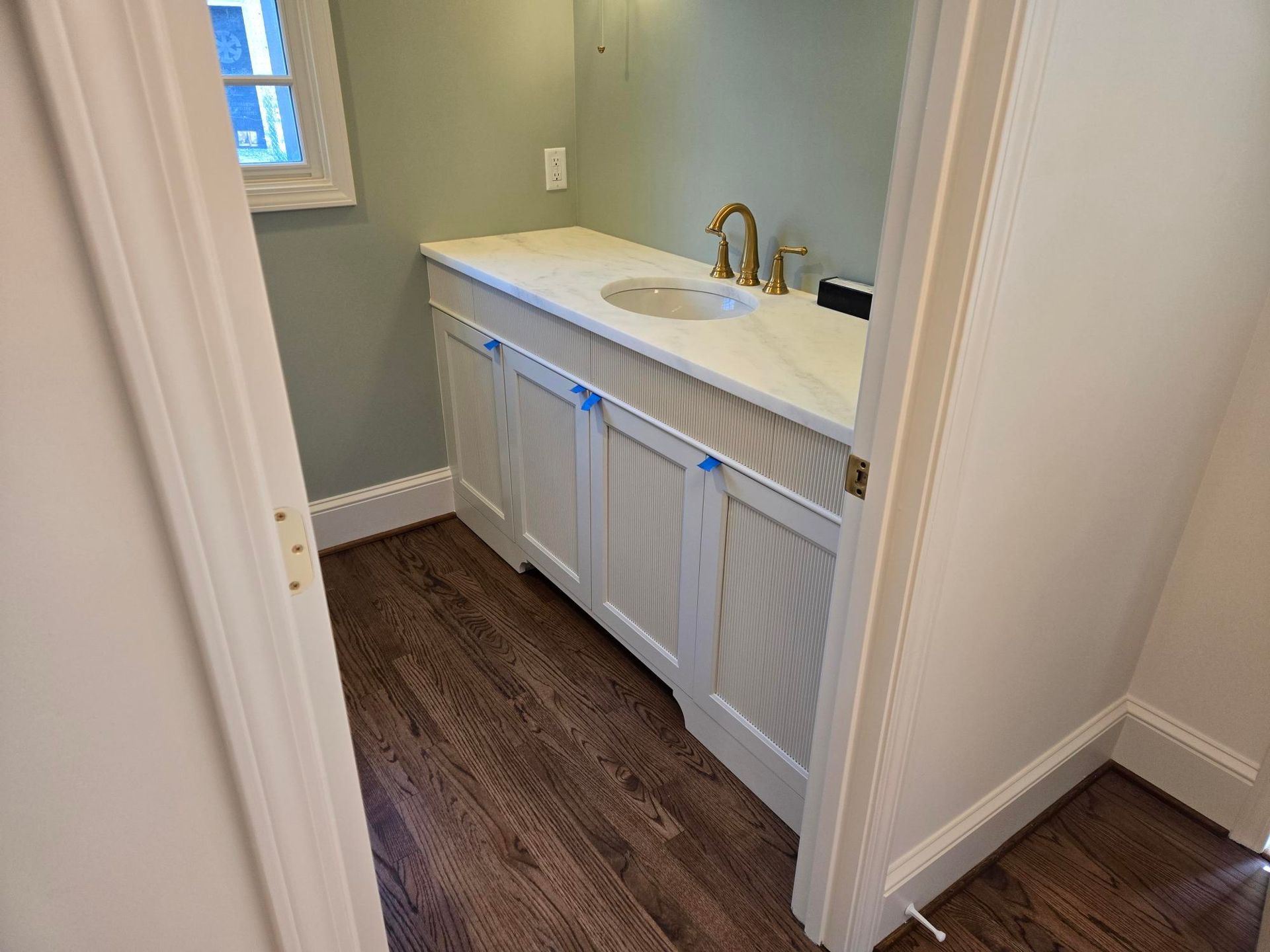 Small bathroom vanity with white cabinets, marble countertop, brass faucet, and dark wood-look floor.