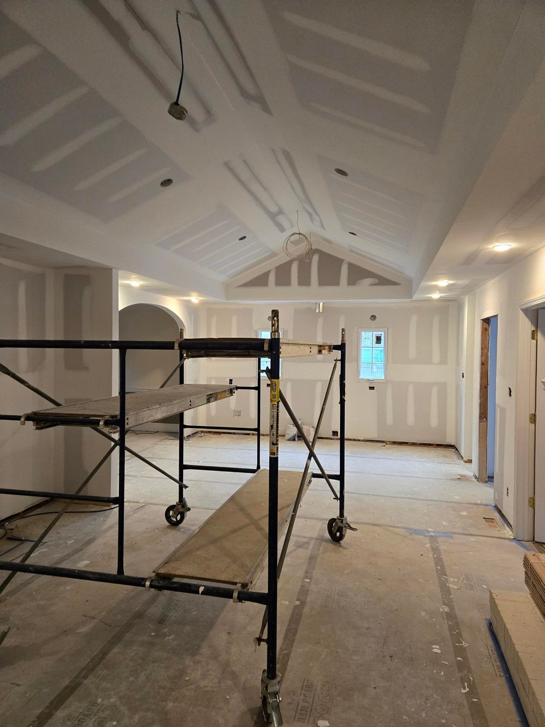 Scaffold setup in a bright, unfinished room with white vaulted ceiling and exposed drywall.