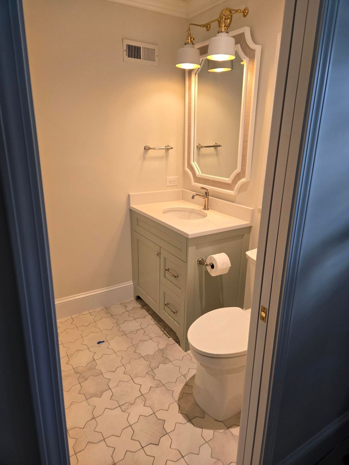 Small bathroom with white vanity, ornate mirror, toilet, and patterned tile floor.
