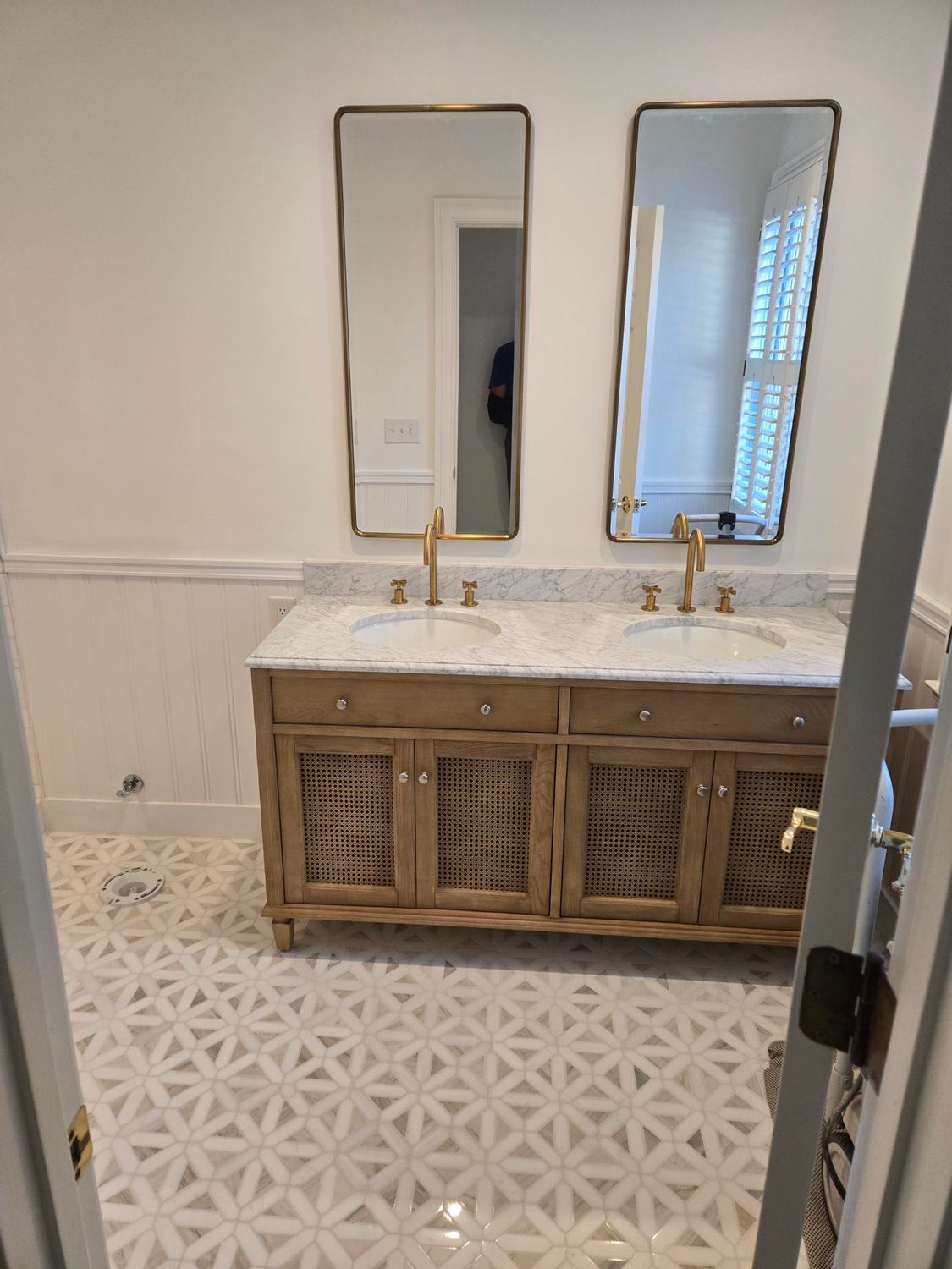Bathroom vanity with marble countertop, two gold faucets, mirrored cabinets, and patterned tile floor.