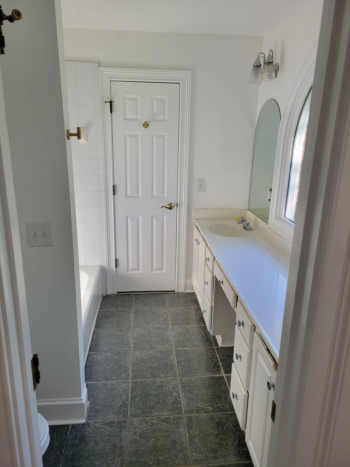 Narrow bathroom with white vanity, round mirror, and dark tile floor facing a closed door.
