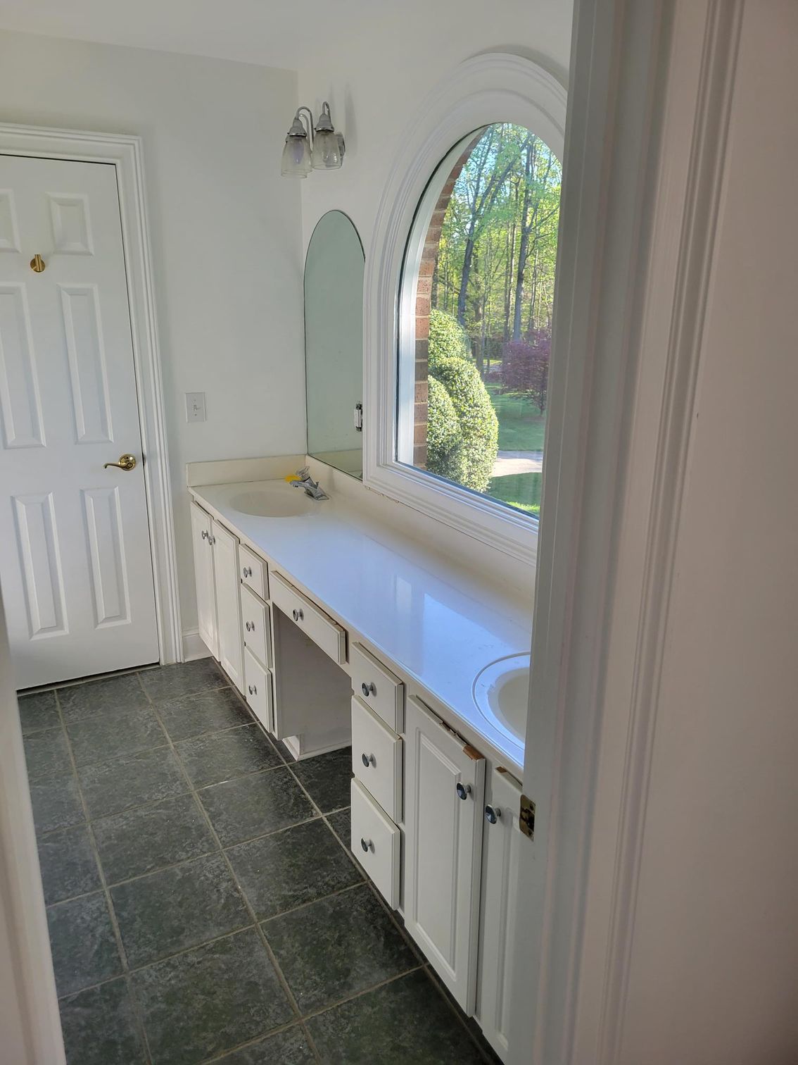 White entryway with built-in cabinet, round mirror, dark tile floor, and arched window overlooking greenery