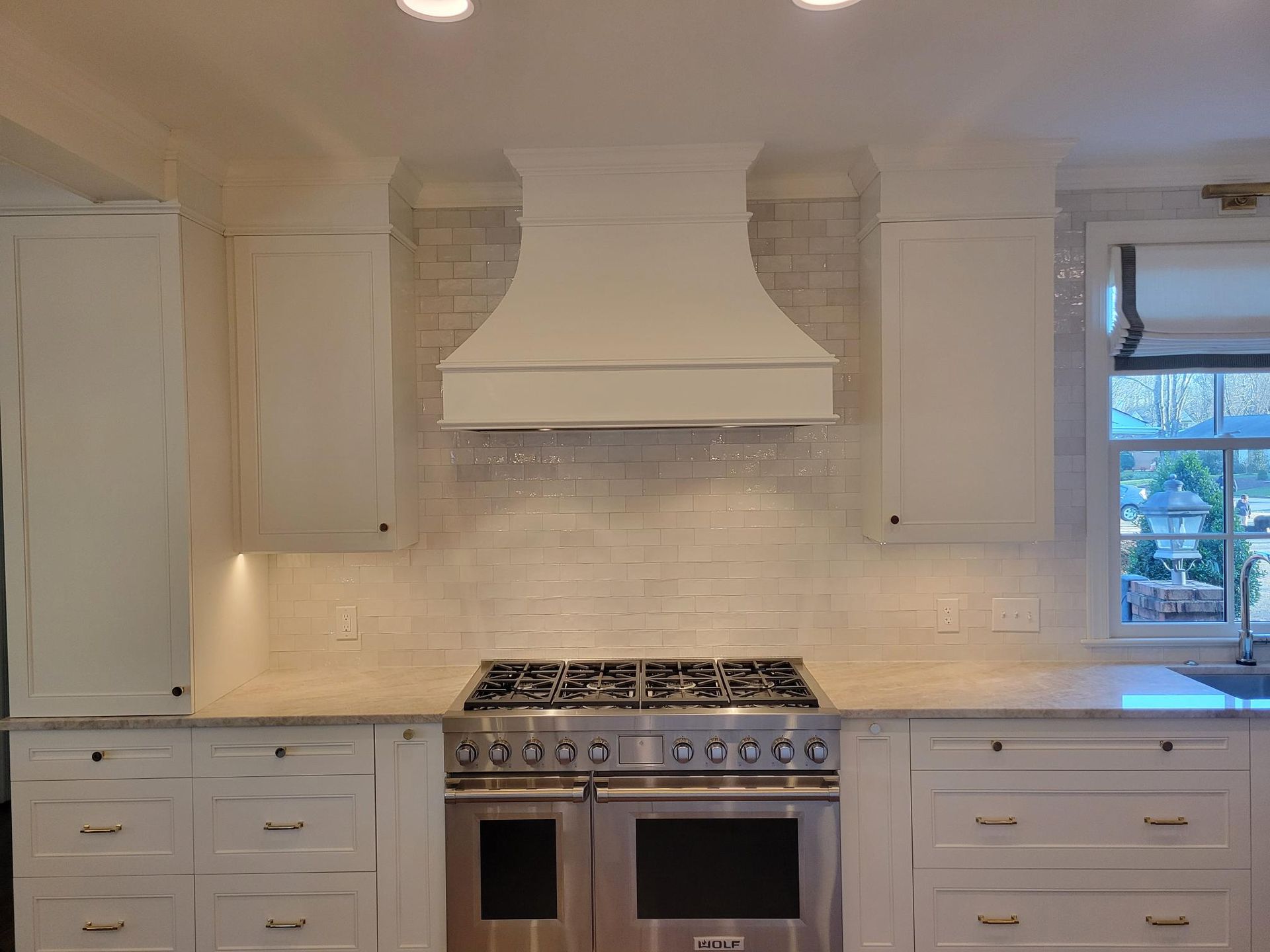 White kitchen with farmhouse hood, marble backsplash, stainless range, and window above counter.