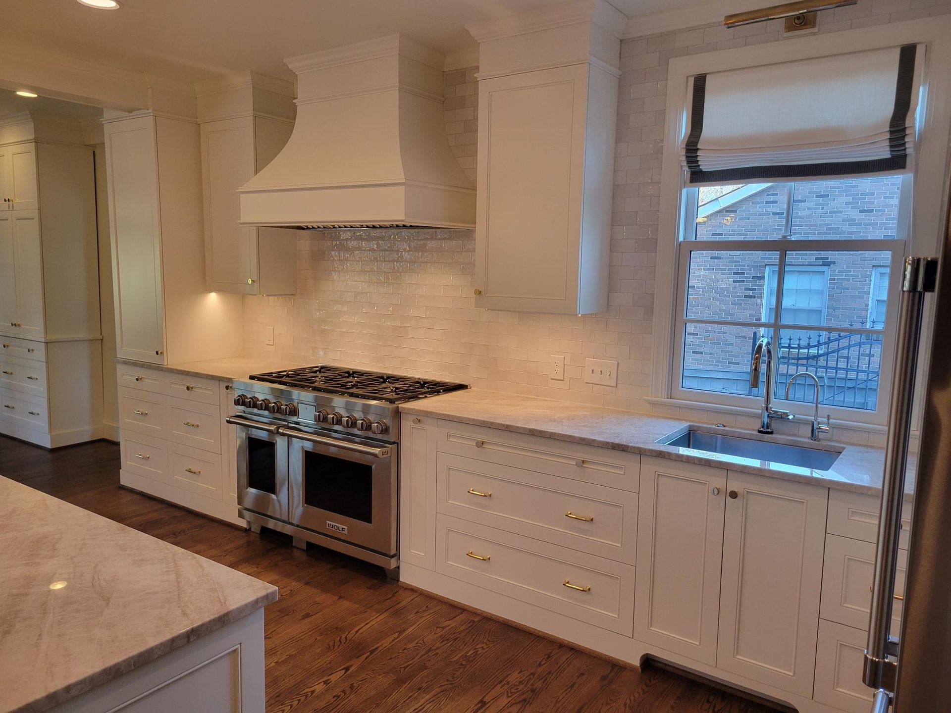Bright white kitchen with wood floors, marble island, stainless stove, and window above the sink.