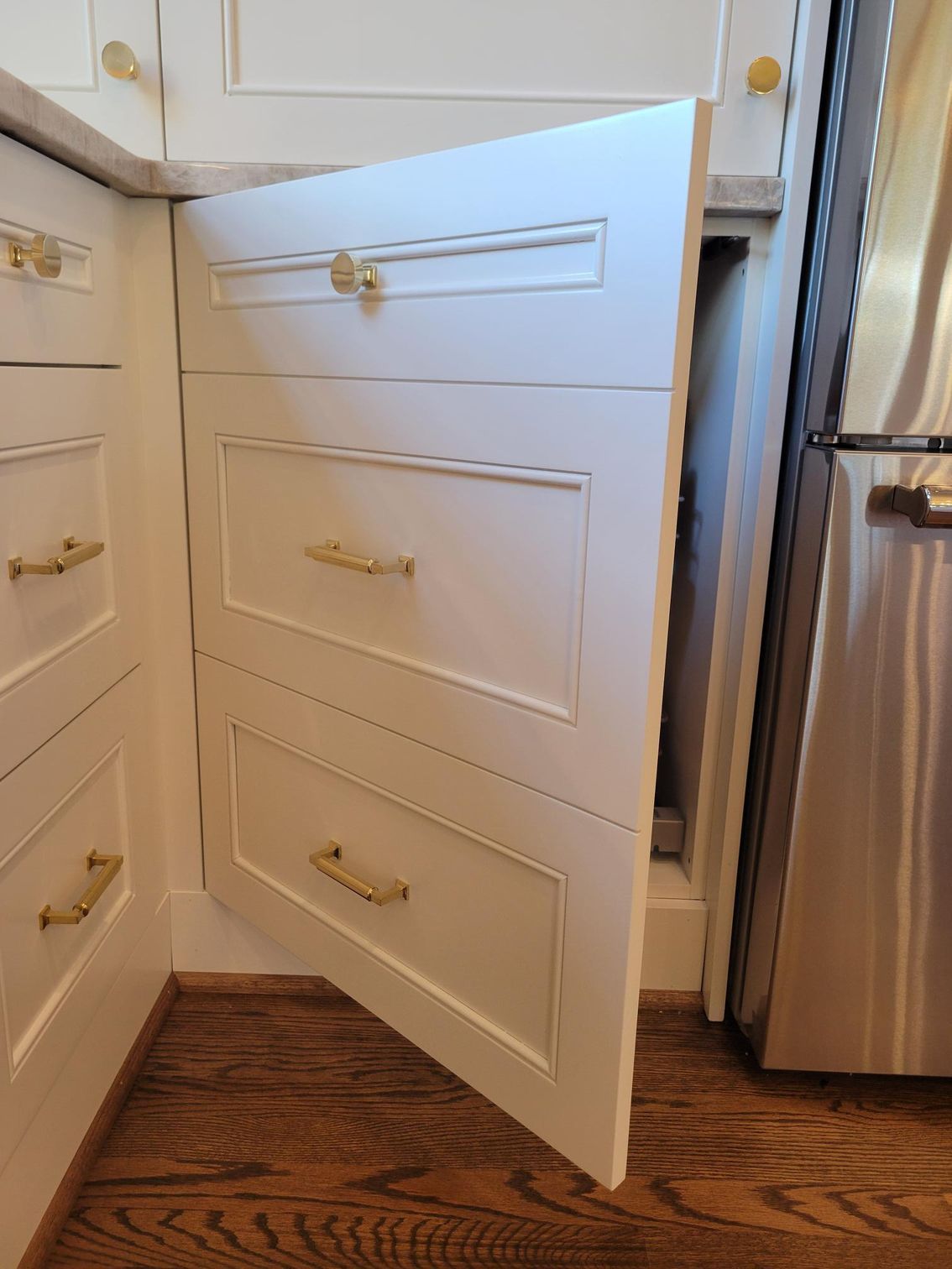 Open white kitchen cabinet with three pull-out drawers beside a stainless steel refrigerator and wood floor.