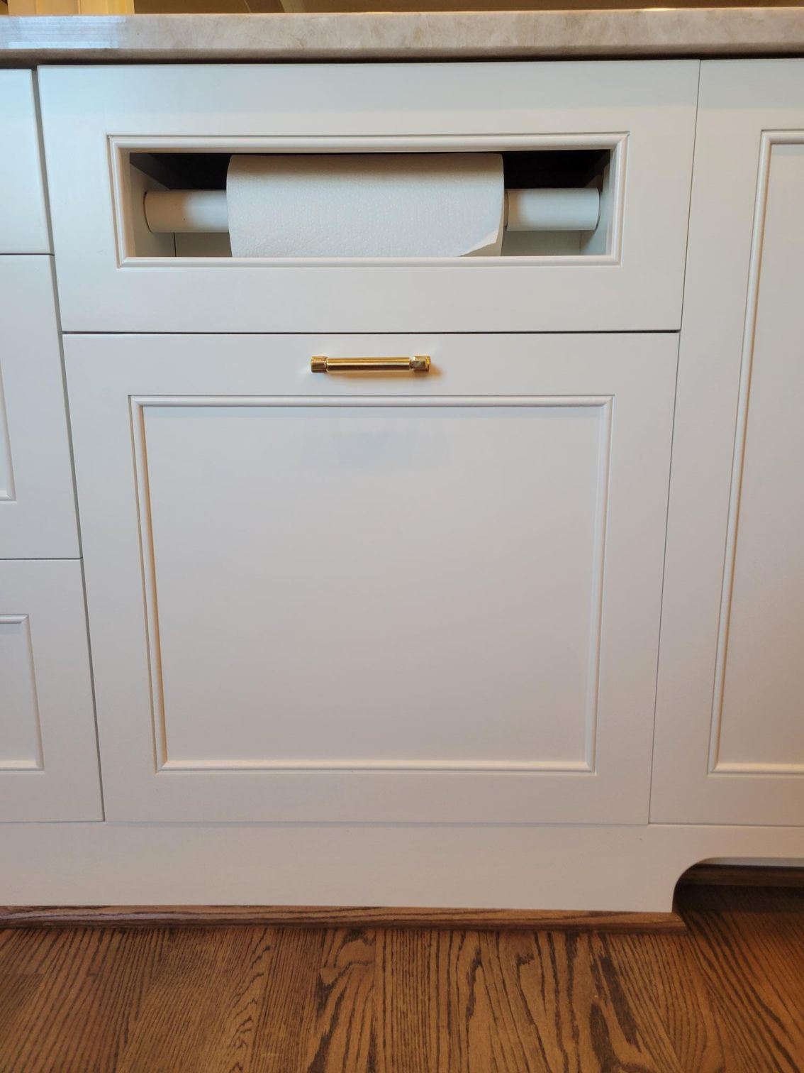 White built-in cabinet with a pull-out paper towel holder and brass handle above hardwood floor.