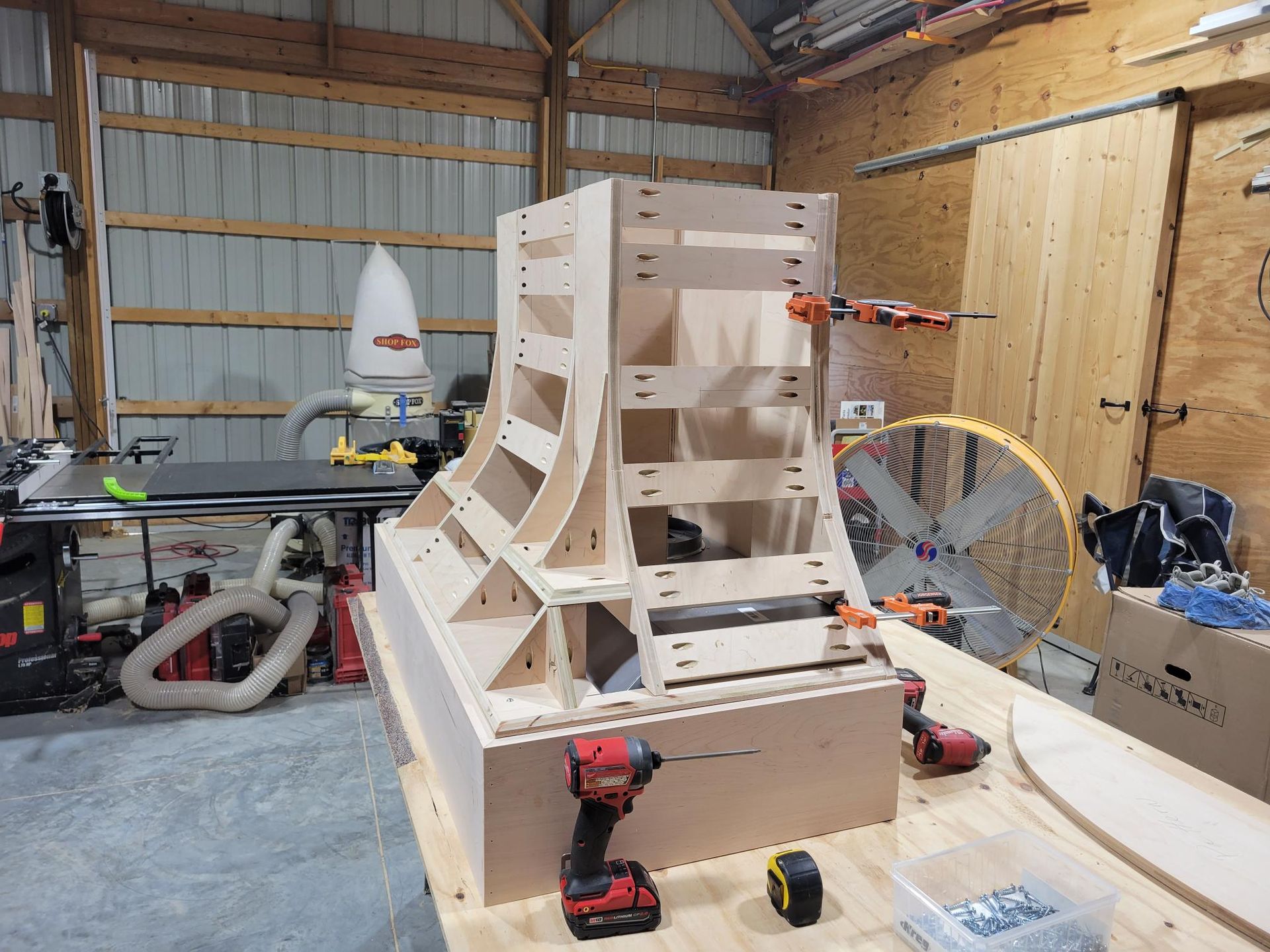 Workshop with a large unfinished wooden machine frame, red clamp, tools, and a fan in the background.