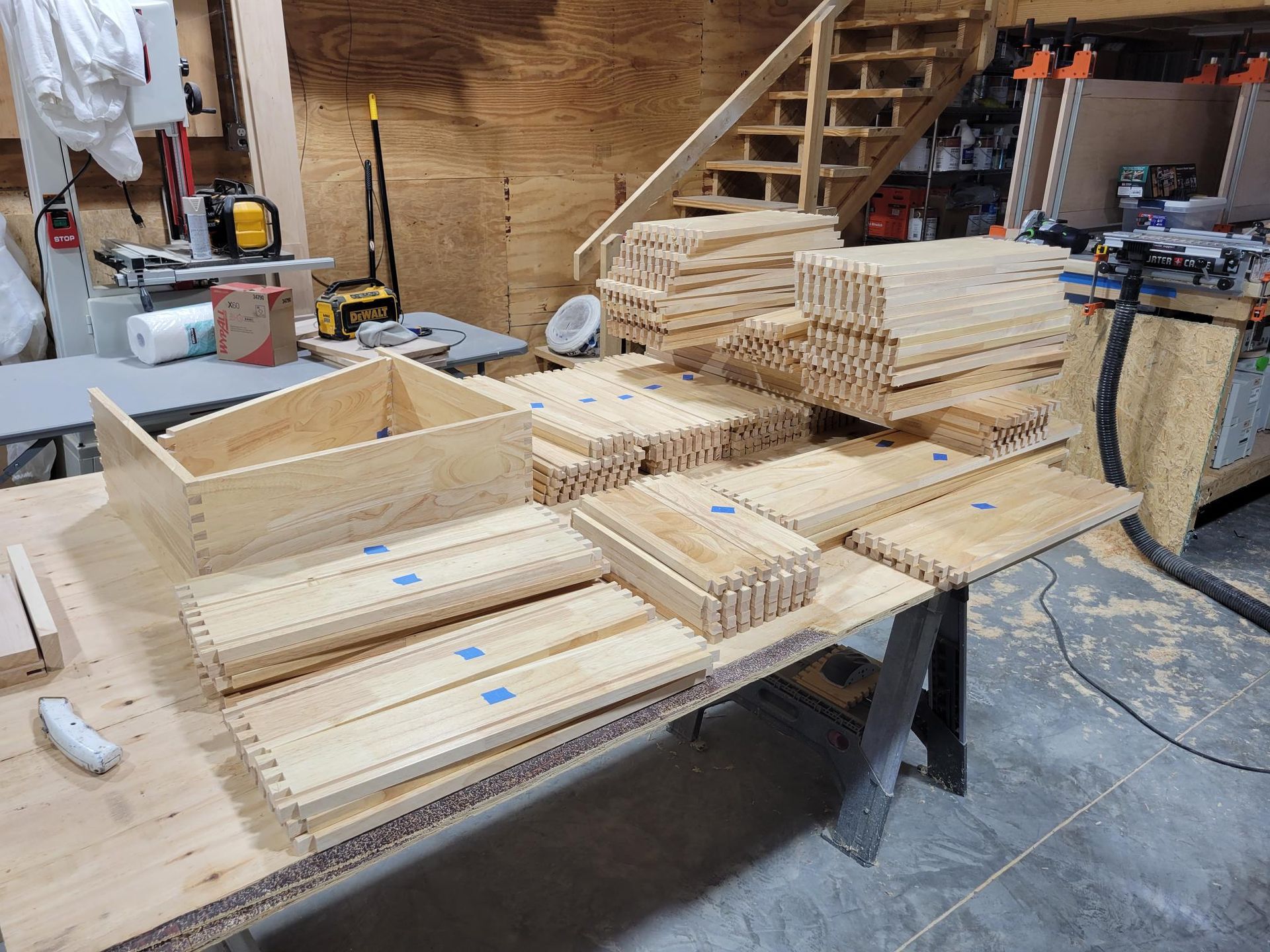 Woodworking shop with stacked wooden drawers, panels, and a staircase in the background.