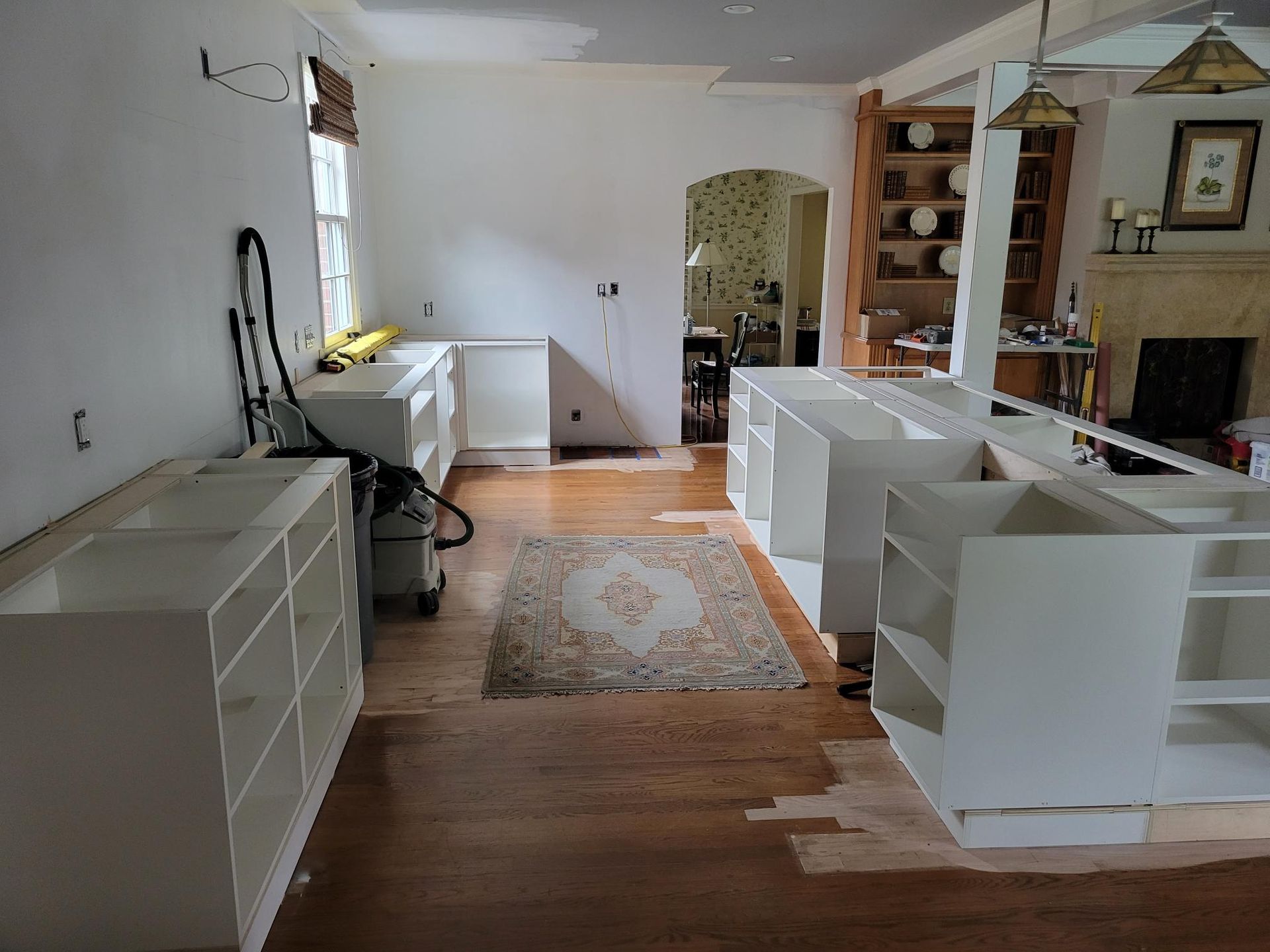 Empty living room with white shelving, a desk by the window, hardwood floors, and a patterned rug.