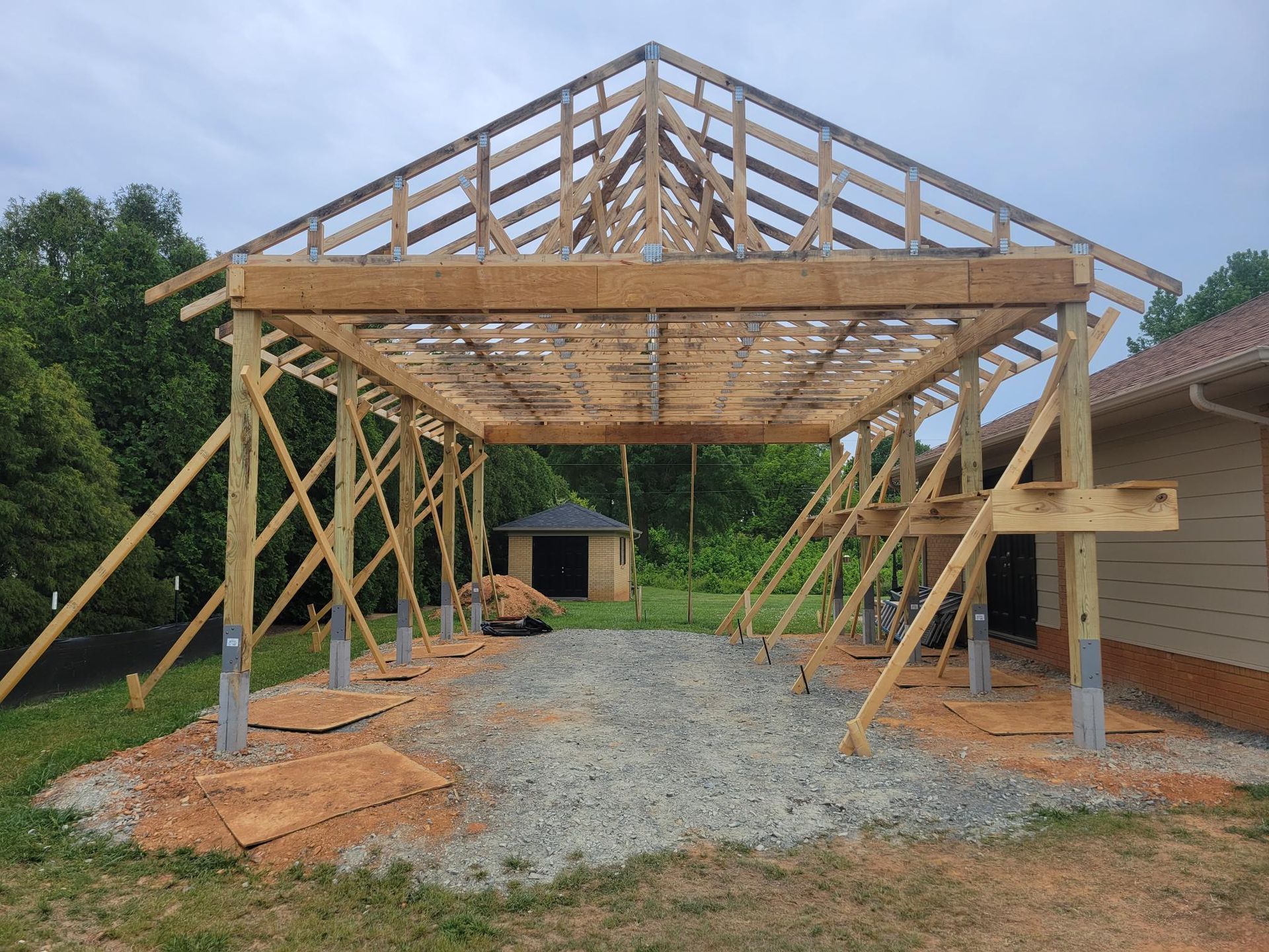 Wooden roof truss framing under construction over a gravel driveway, with support posts and braces.
