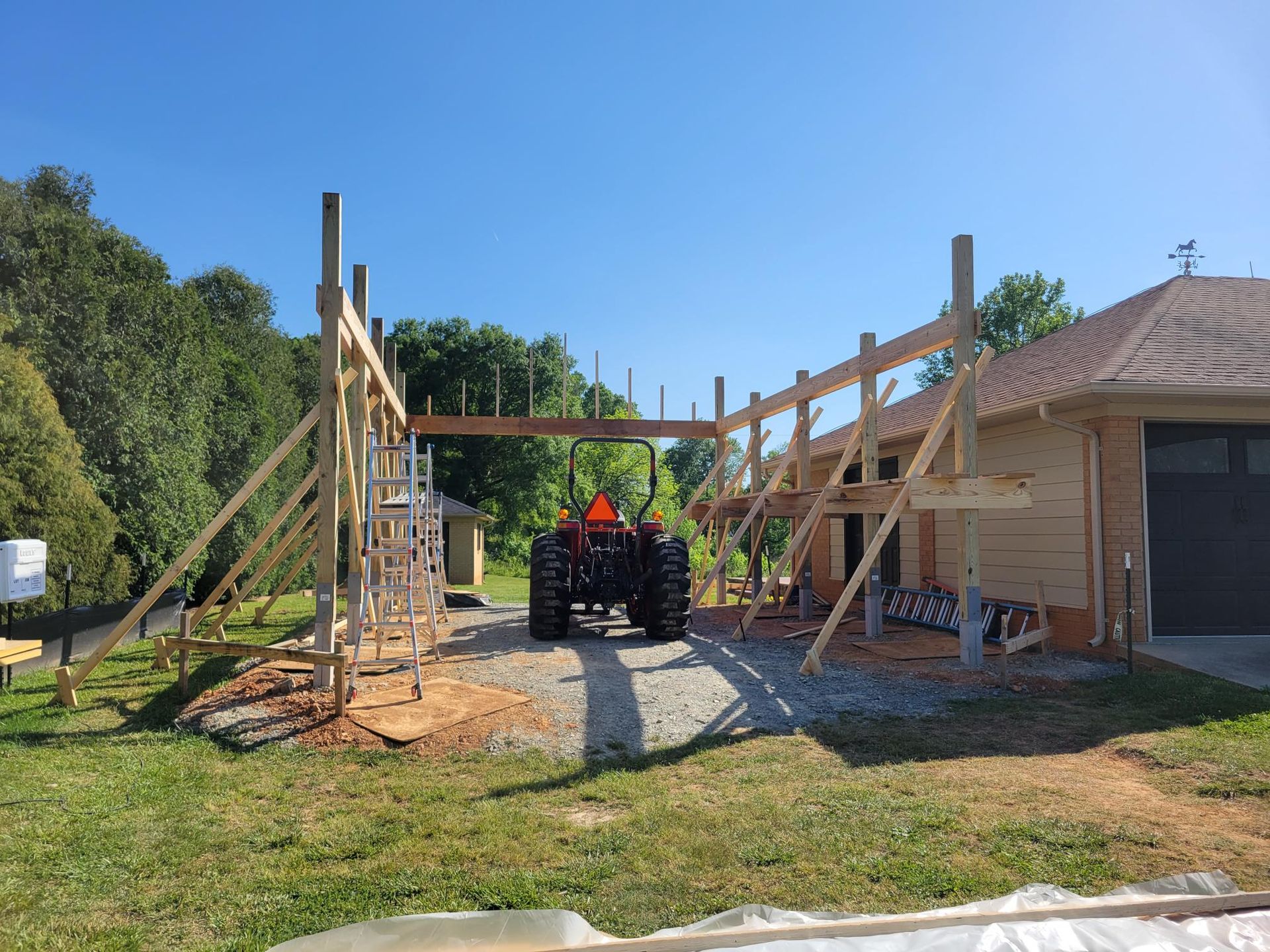 Construction site with wooden framing under a clear blue sky beside a house and tractor.
