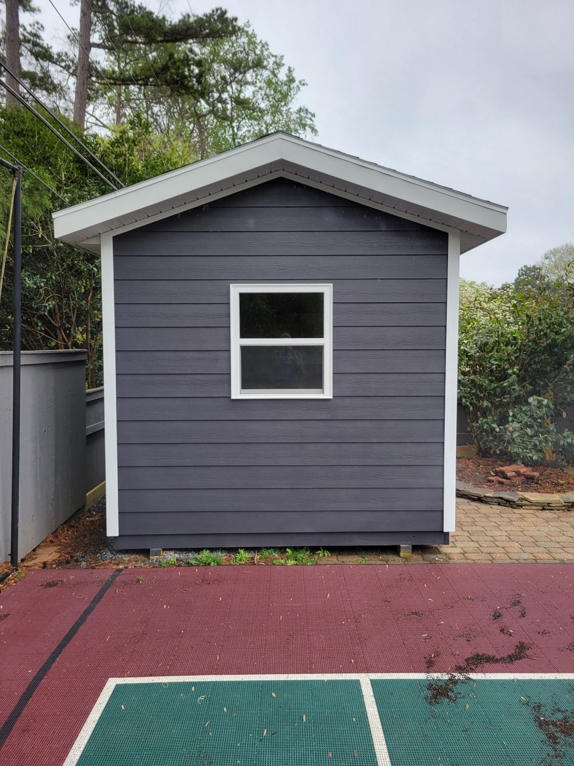 Small gray shed with a single window beside a red paved court and trees outdoors