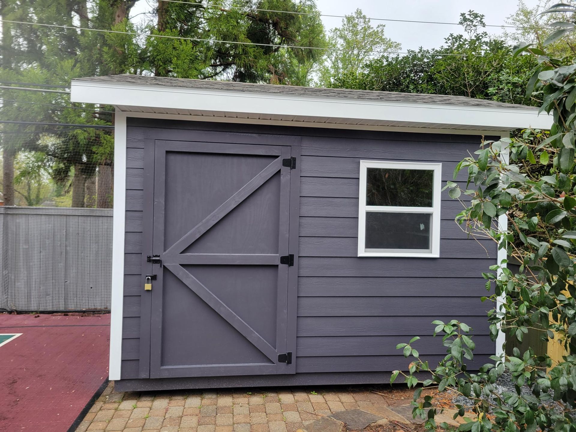 Gray backyard shed with a white window and slanted roof next to a red patio and fence