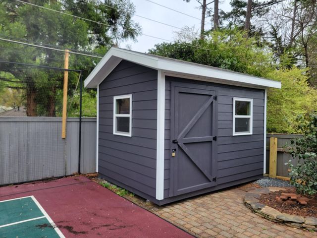 Small gray backyard shed with white trim beside a fence and paved patio area