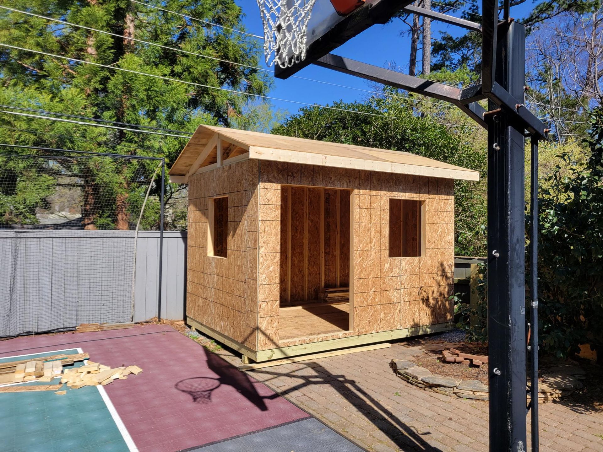 Small unfinished wooden shed in a backyard beside a basketball hoop and fenced yard.