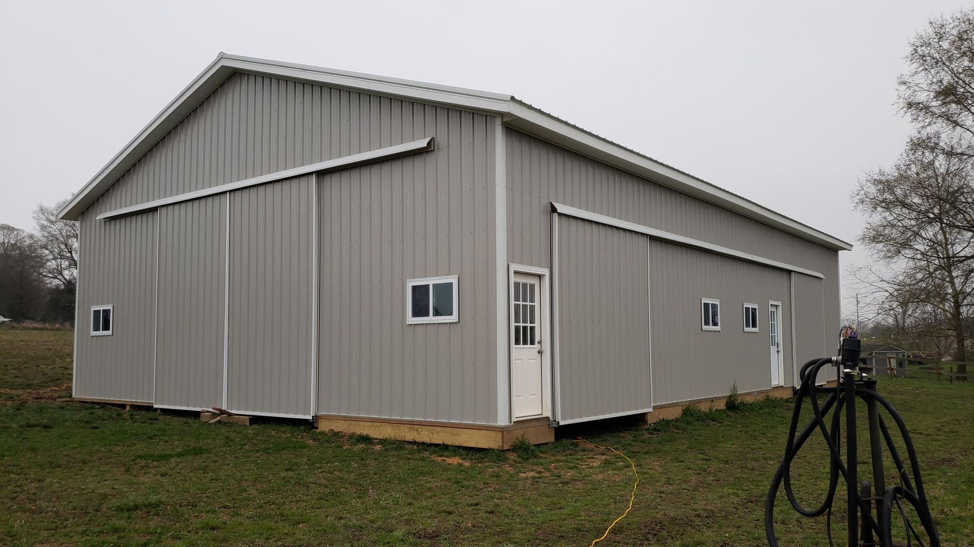 Gray metal barn-like building with a gabled roof in a grassy field under an overcast sky