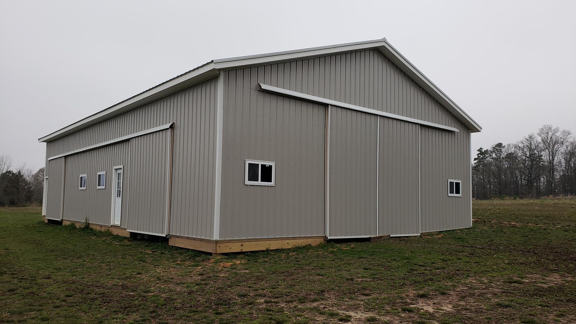 Large gray metal barn with multiple doors and windows on a grassy field under an overcast sky