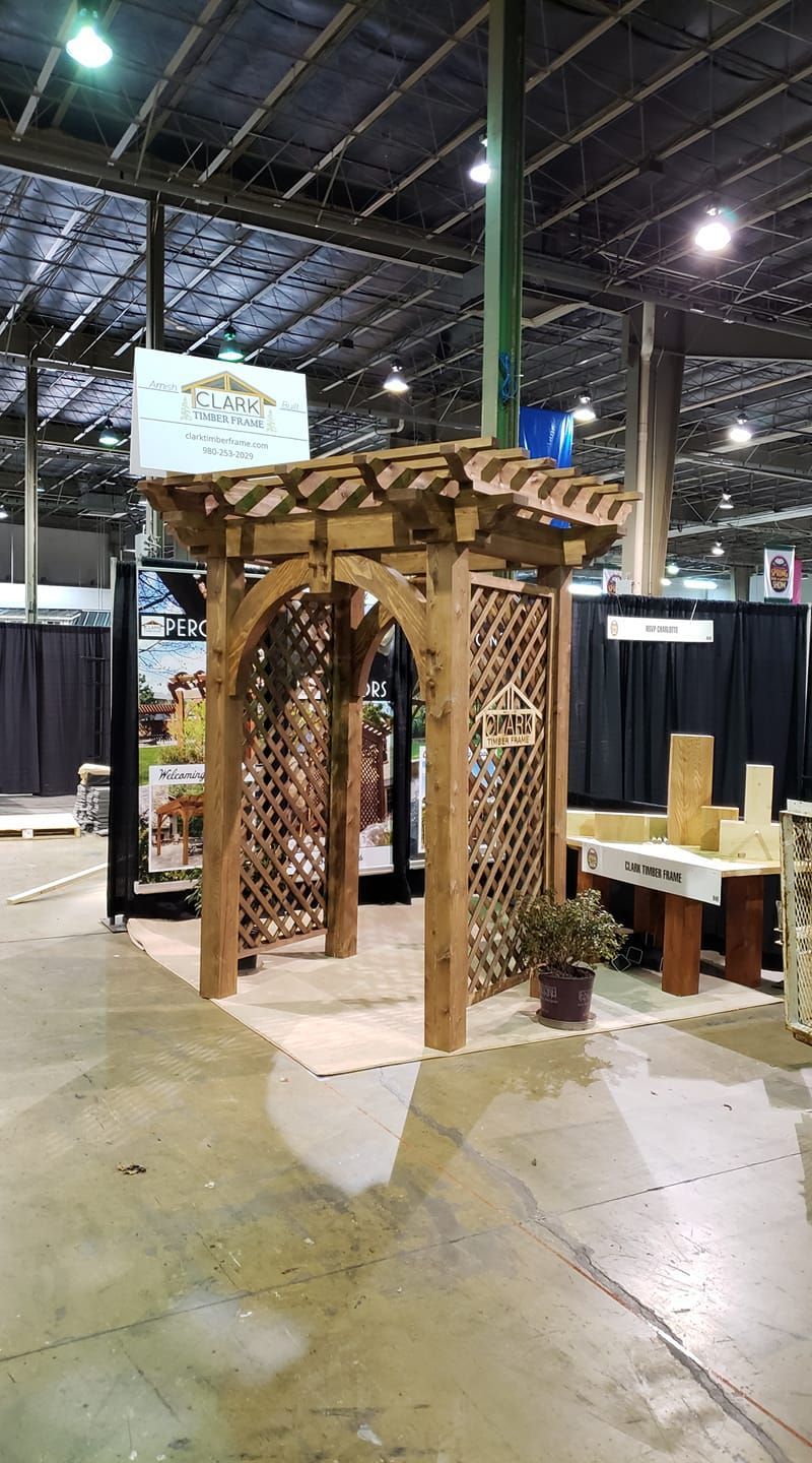 Wooden pergola-style booth display at an indoor trade show, with a sign and chairs nearby.